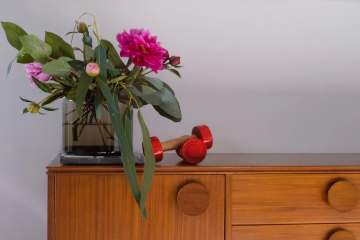 Pfingstrosen in einer Glasvase auf einem Teakholz-Sideboard mit runden Griffen, daneben rote Hanteln