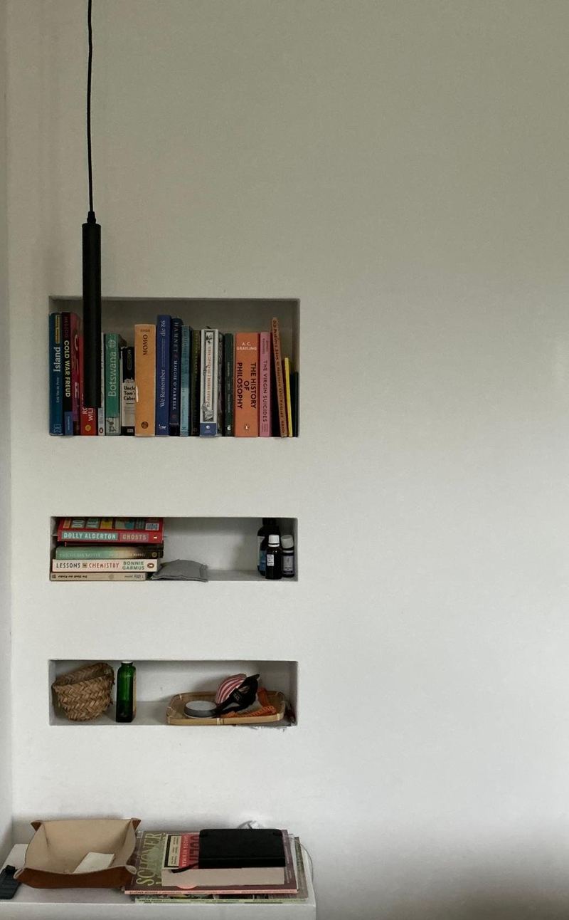Weiße Wand mit drei eingelassenen Nischen voller Bücher, davor eine schwarze Pendelleuchte
